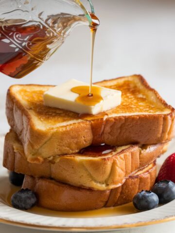 A stack of Brioche French Toast Recipe topped with a pat of butter and drizzled with warm maple syrup. The thick, golden-brown slices are crisp on the outside and soft on the inside, served on a classic plate with fresh blueberries and a ripe strawberry. This French toast recipe is a rich, indulgent breakfast treat perfect for any morning!