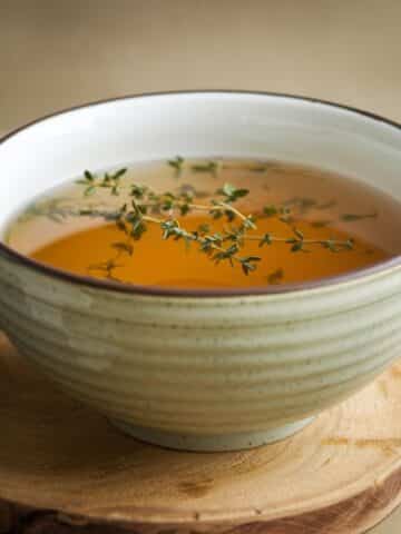 A rustic ceramic bowl filled with golden homemade chicken broth Recipe, garnished with fresh thyme sprigs, placed on a wooden cutting board. The broth has a rich, clear texture, making it perfect for soups, stews, or sipping