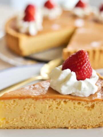 A slice of golden, creamy Impossible Pie topped with whipped cream and a fresh strawberry, served on a white plate with a full pie in the background. This easy, self-crusting Impossible Pie Recipe is a delicious and classic dessert perfect for any occasion.