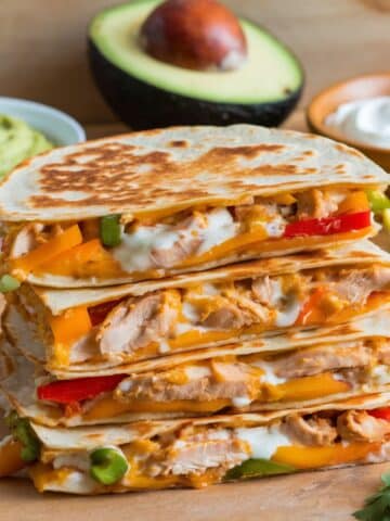 Stacked chicken quesadillas filled with melted cheese, juicy chicken, and colorful bell peppers, served with guacamole, sour cream, and fresh avocado in the background, perfect for a Chicken Quesadilla Recipe.