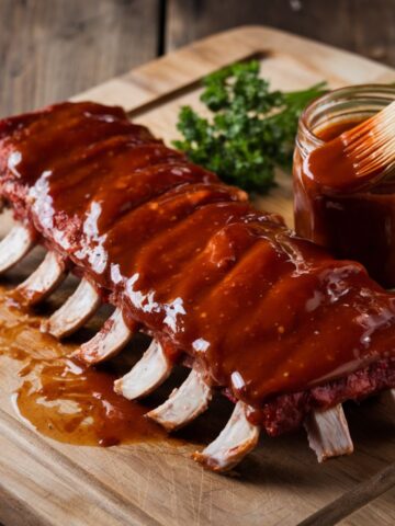 A rack of beef back ribs generously coated in a thick, tangy barbecue sauce, resting on a wooden cutting board. A basting brush and jar of sauce are nearby, adding extra flavor. Perfect for grilling, smoking, or slow cooking, this beef back ribs recipe delivers juicy, fall-off-the-bone tenderness.