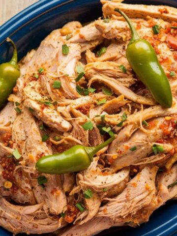 Shredded Mississippi Chicken Recipe served on a blue platter, topped with tangy pepperoncini peppers and fresh herbs for a juicy, flavorful slow-cooked dish.