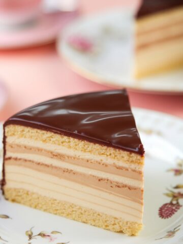 A slice of Boston Cream Cake featuring layers of fluffy vanilla sponge cake, rich custard filling, and a smooth, glossy chocolate ganache topping, served on an elegant floral plate. This classic dessert is indulgent and perfect for any occasion.