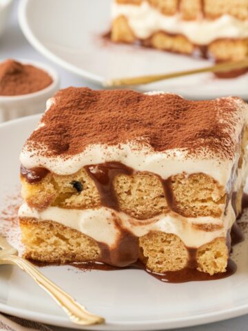 A homemade slice of gluten free tiramisu served on a white plate with a gold fork beside it. The dessert consists of two layers of coffee-soaked gluten-free ladyfingers, enveloped in a rich mascarpone cream filling. The mascarpone mixture is slightly melting over the edges, creating an irresistible, creamy texture.