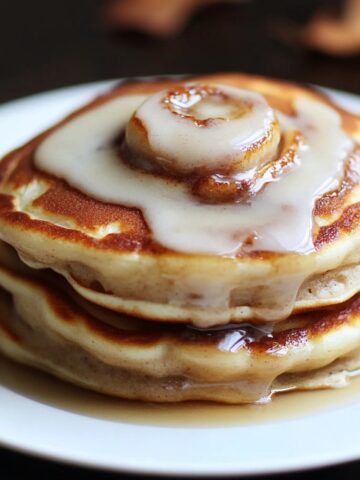 Homemade cinnamon roll pancakes stacked on a white plate, topped with creamy cinnamon glaze and maple syrup drizzle.