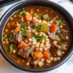 Homemade beef barley soup with tender beef, carrots, celery, and pearl barley in a rich broth, topped with fresh parsley in a black bowl.