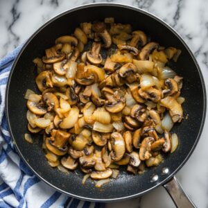 Sautéed mushrooms and onions in a black skillet, sitting on a marble counter with a blue-striped kitchen towel-part of the filling for beef Wellington.