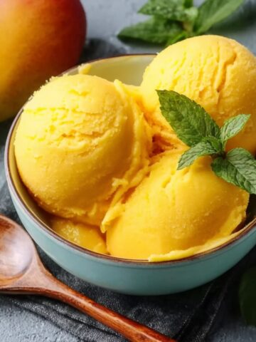Three scoops of bright yellow mango sorbet in a teal bowl, topped with fresh mint, next to a wooden spoon and a ripe mango.