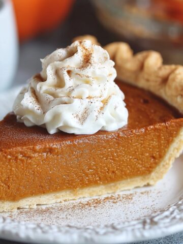 A slice of homemade pumpkin pie with a golden, flaky crust and creamy orange filling, topped with whipped cream and cinnamon, on a white plate with coffee and pumpkins in the background.