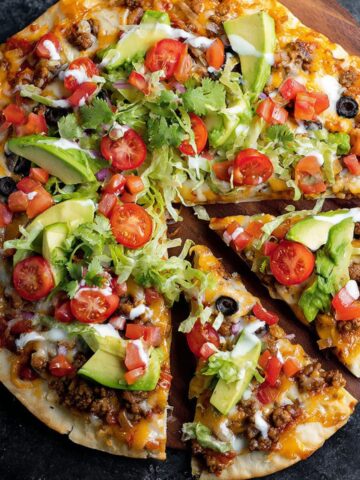 Homemade taco pizza topped with ground beef, cheese, lettuce, tomatoes, avocado, and sour cream on a wooden board.
