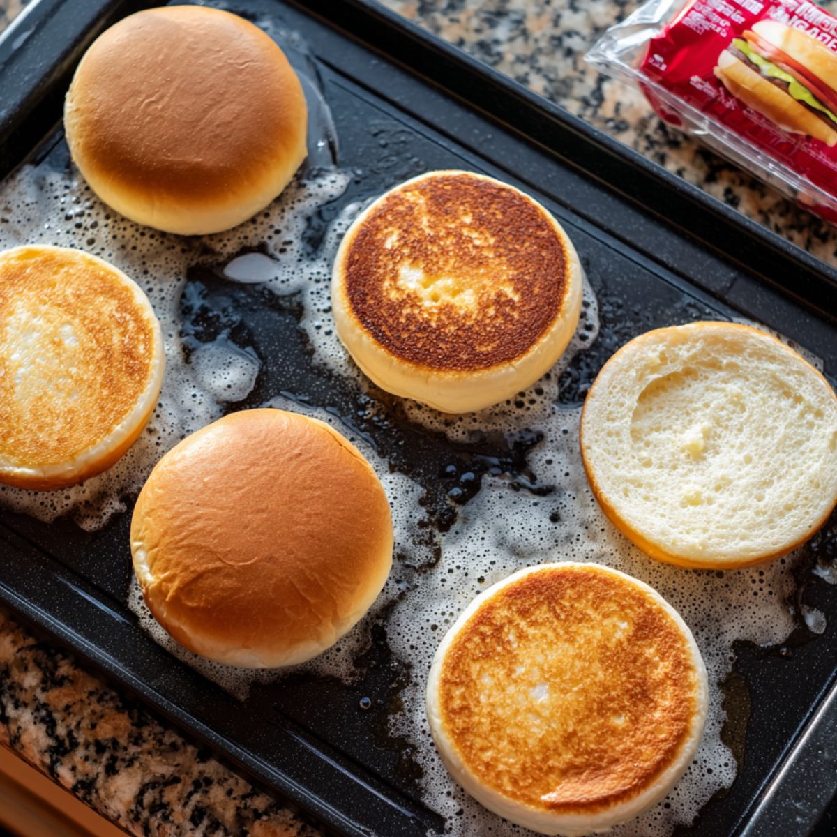 Top-down view of six hamburger buns toasting on a black griddle, with melted butter bubbling around them. Some buns are golden brown on the cut side, others show their soft tops. A bun package is visible on a granite countertop, giving a homemade, real-kitchen feel.