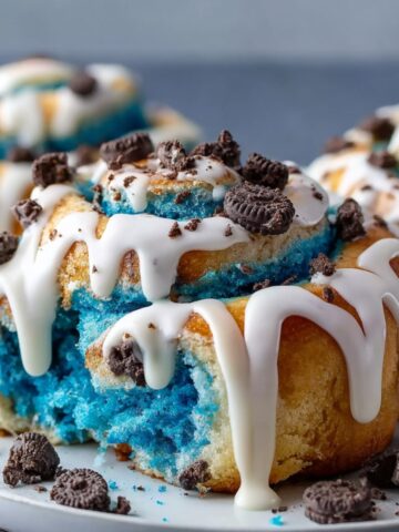 Homemade Cookie Monster cinnamon rolls with blue swirled dough, white icing drizzle, Oreo crumbles, and golden edges.