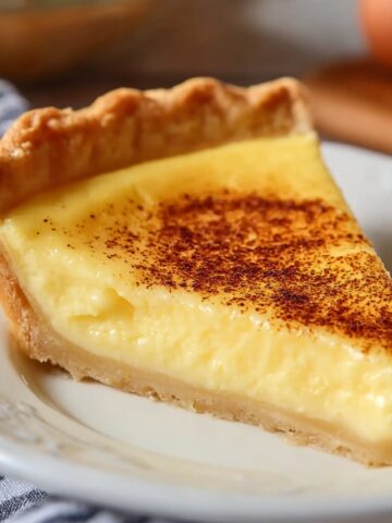 Close-up of a homemade egg custard pie slice on a vintage white and gray patterned plate, showing a smooth, creamy yellow custard filling topped with a golden-brown nutmeg-speckled surface and a flaky, buttery pie crust edge, softly lit on a wooden table with a blurred background for a warm, inviting kitchen feel.