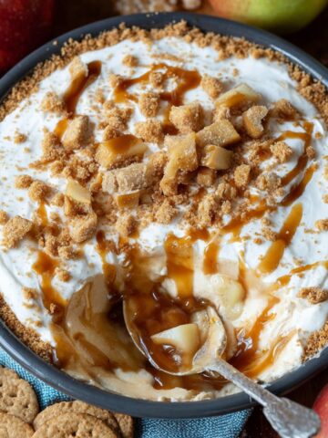 Homemade caramel apple cheesecake dip topped with caramel drizzle, crumbly brown sugar streusel, and diced apples in a round dish, surrounded by apples and graham crackers.