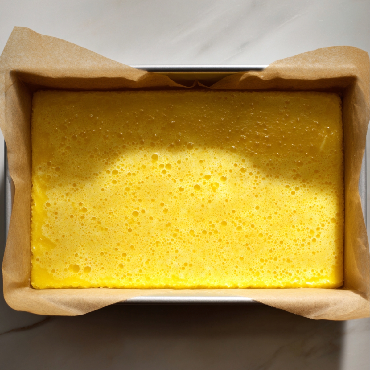 Freshly baked homemade lemon bars in a parchment-lined pan, with a bright yellow, slightly bubbly surface under natural sunlight.