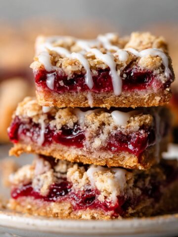 A stack of freshly baked Cherry Pie Bars with a golden, crumbly oat topping, filled with rich cherry filling and topped with a light drizzle of icing.