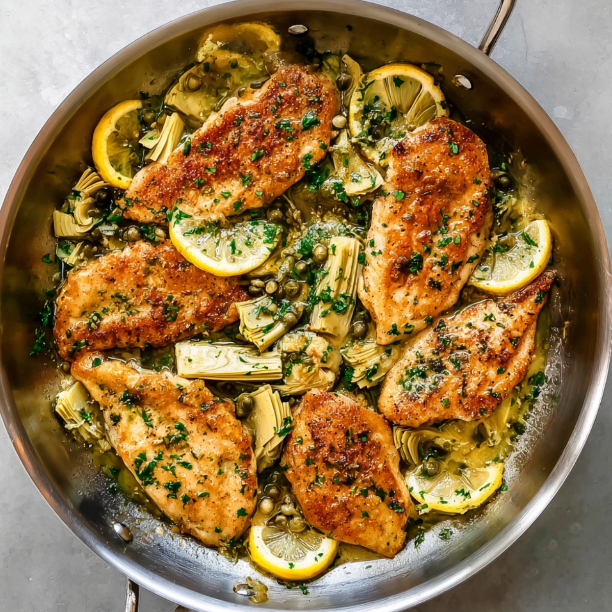 Lemon artichoke chicken in a skillet with golden seared chicken breasts, artichoke hearts, capers, parsley, and lemon slices in a buttery sauce.