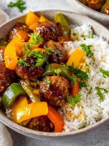 Sweet and sour meatballs served with colorful peppers, garnished with fresh parsley on a bed of rice.