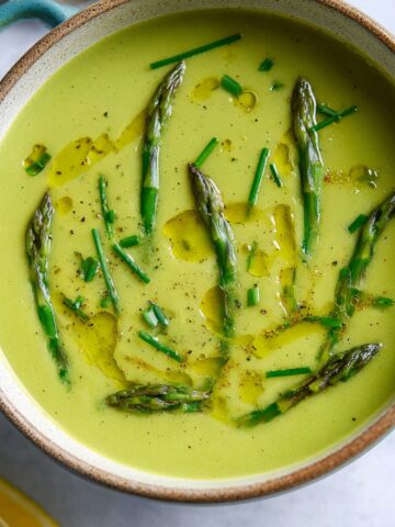 A bowl of creamy Asparagus Soup, garnished with whole asparagus spears, a drizzle of olive oil, and fresh chives, creating a smooth and vibrant dish.