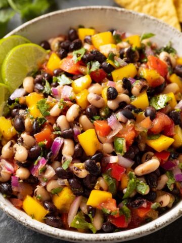 A bowl of Cowboy Caviar Recipe, featuring black beans, black-eyed peas, tomatoes, yellow bell peppers, red onions, and cilantro, garnished with lime wedges.