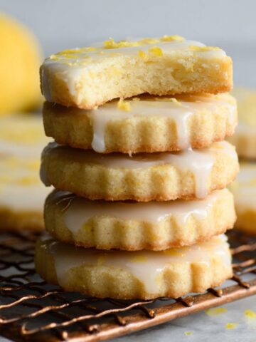 A stack of Lemon Shortbread Cookies with a glaze and lemon zest topping, one cookie with a bite taken out, freshly baked and cooling on a wire rack.