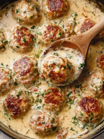 A close-up of Creamy Marry Me Chicken Meatballs in a skillet, simmering in a rich sauce, with a wooden spoon lifting one meatball, surrounded by a smooth, savory sauce.