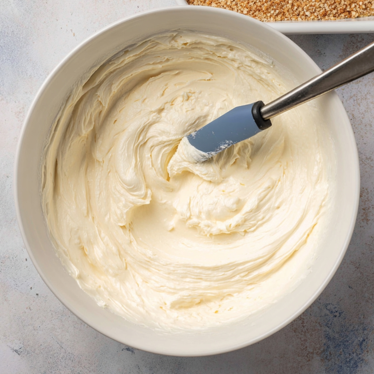 A creamy filling being mixed in a white bowl with a spatula. The texture is smooth and fluffy, ready to be spread onto the pretzel crust for a dessert layer.