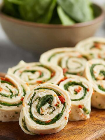 Turkey pinwheels made with tortillas, spinach, and creamy filling, neatly sliced and arranged on a wooden board, with a bowl of fresh spinach in the background.